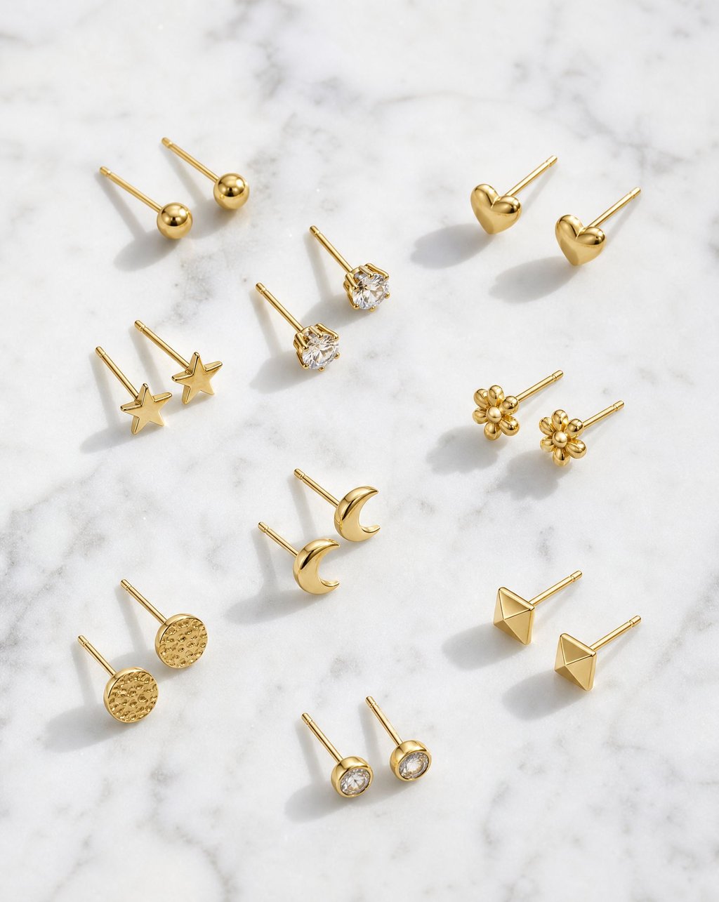 An overhead flat lay of nine pairs of small gold stud earrings on white marble