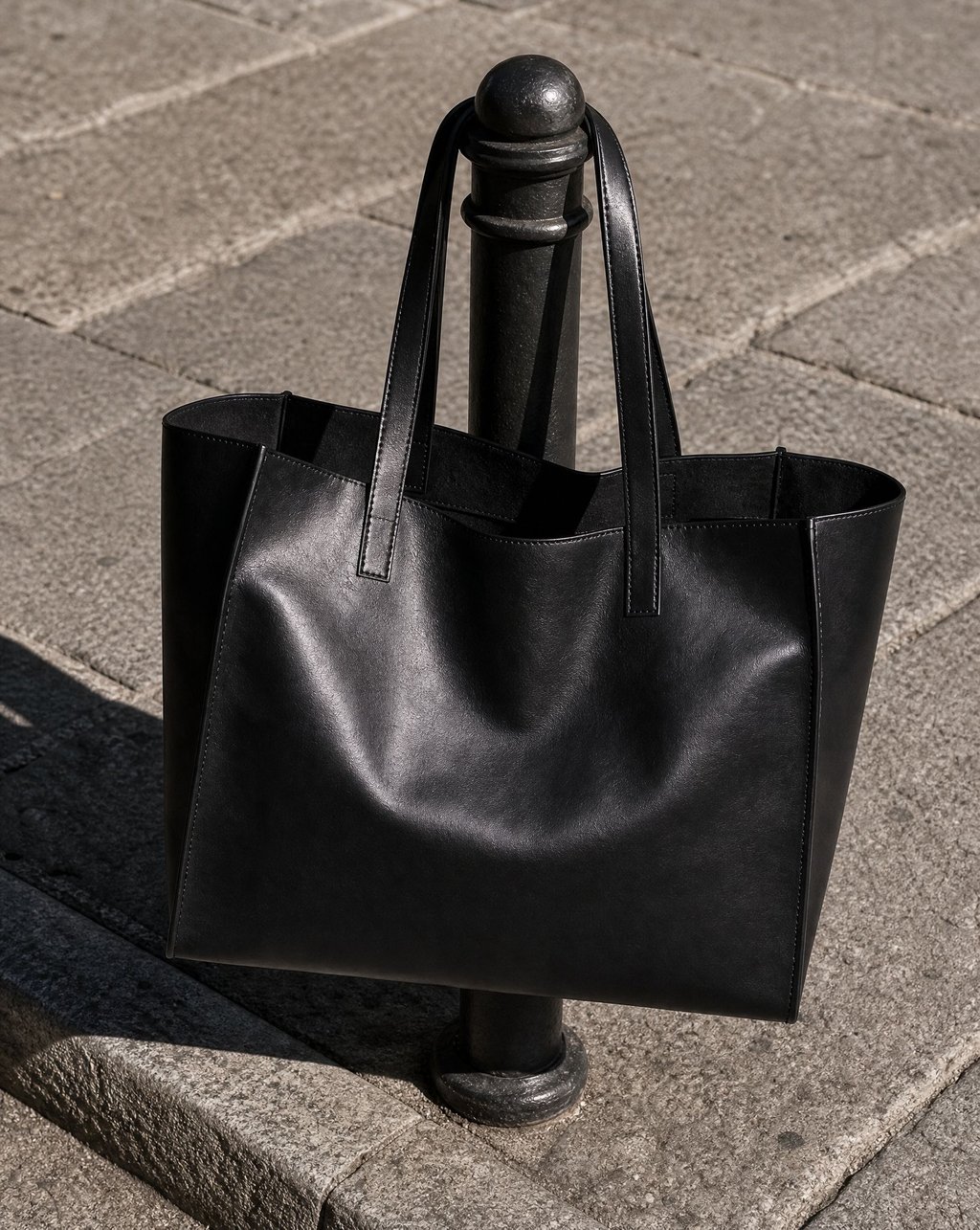 A black leather tote bag hanging by its straps on a cast-iron street bollard in hard sunlight