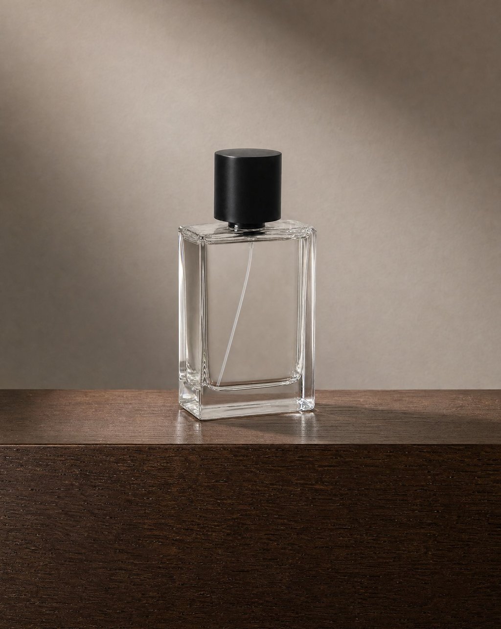 A clear glass perfume bottle on dark timber against a warm plaster wall under a soft pool of light