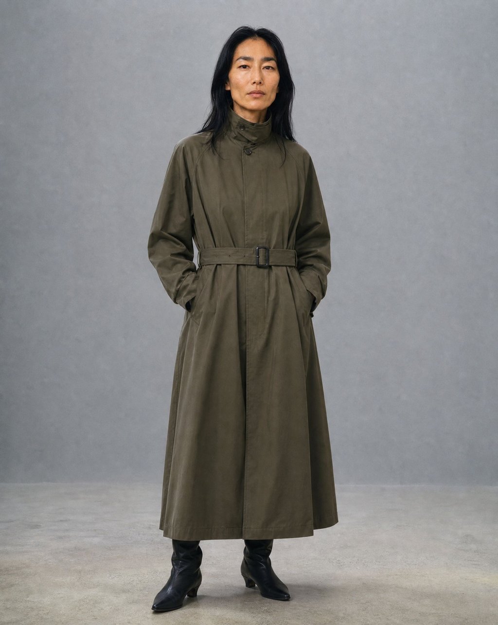A woman with long black hair wearing a belted olive green trench coat against a grey studio wall