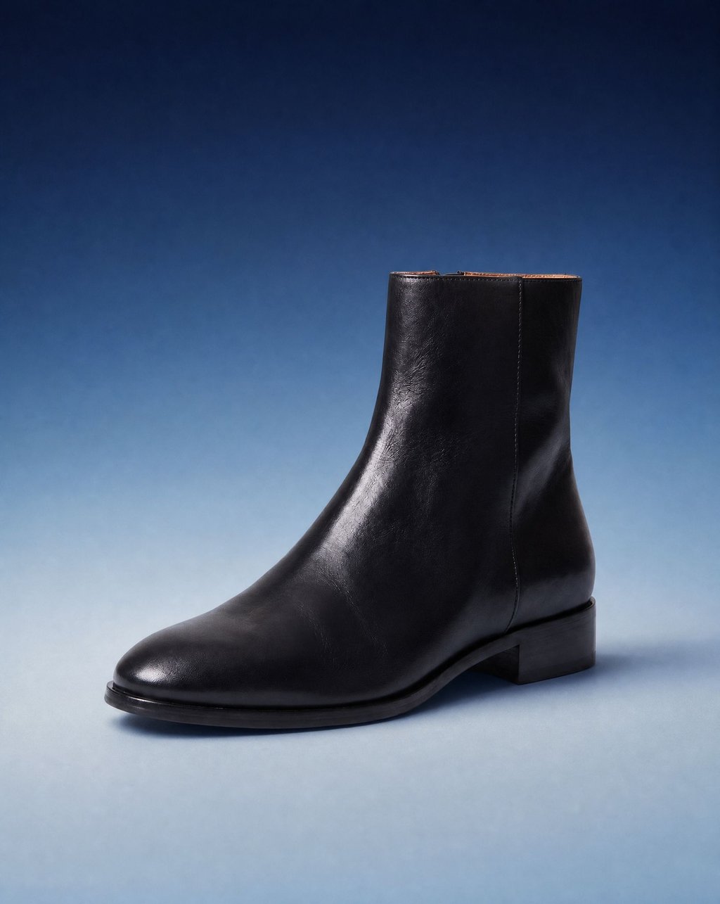 A black leather ankle boot on a blue gradient backdrop under soft even light, with a soft shadow beneath it and no hard shadow edge