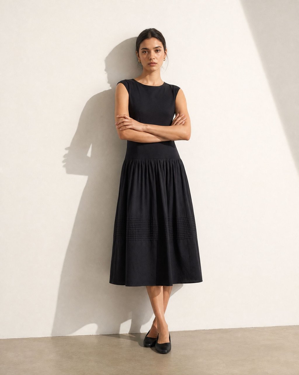 A model in a black pleated midi dress against a pale wall under a strong directional shadow, the pintuck band and hem clearly resolved