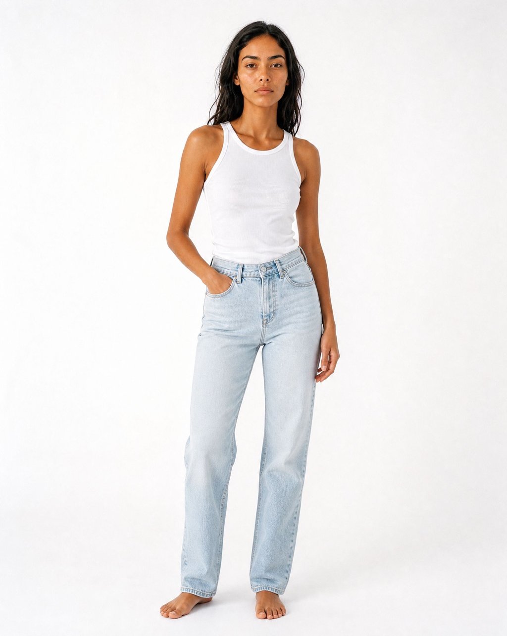 A woman with dark wavy hair wearing the same white ribbed tank top with light jeans in a white studio