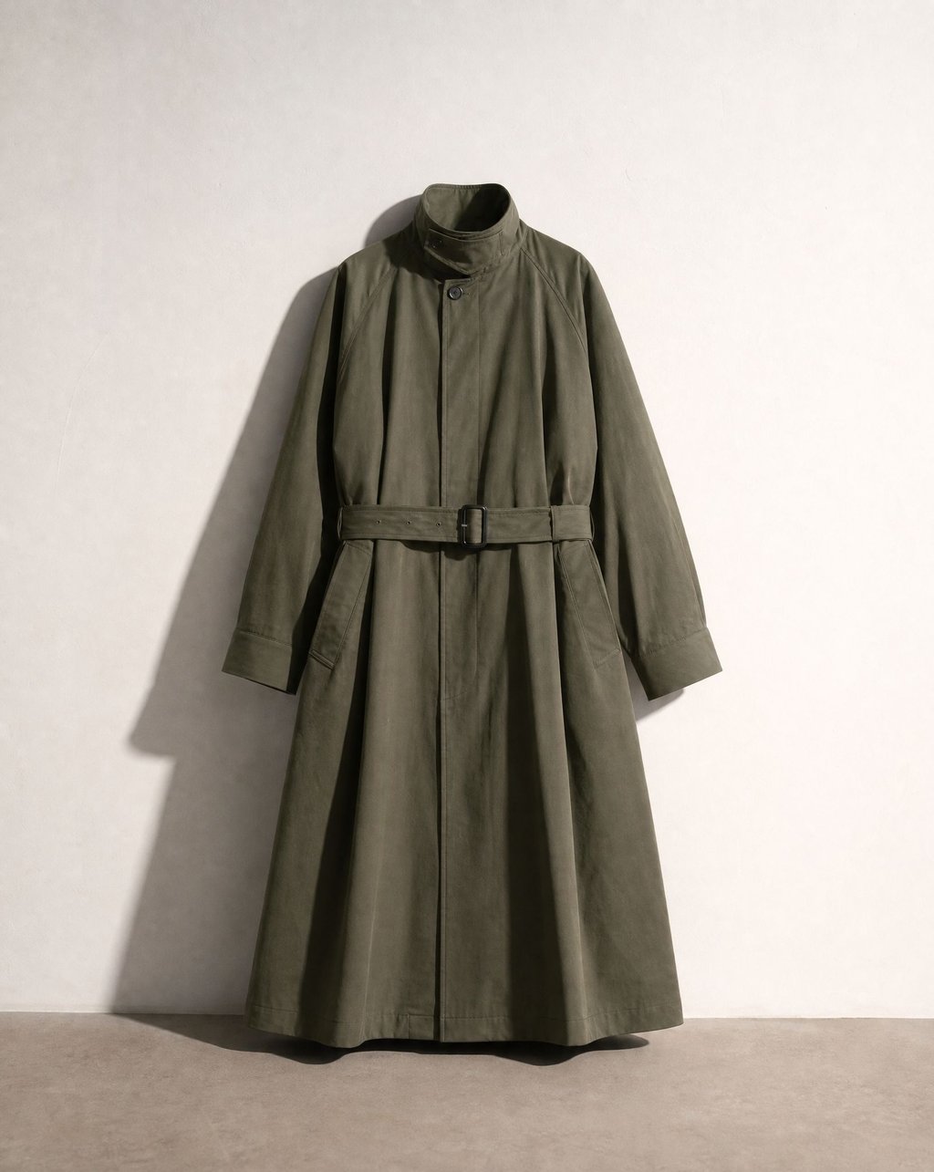 An olive green belted trench coat presented flat against a pale wall with a strong directional shadow falling to the left, the belt buckle and collar structure clearly visible