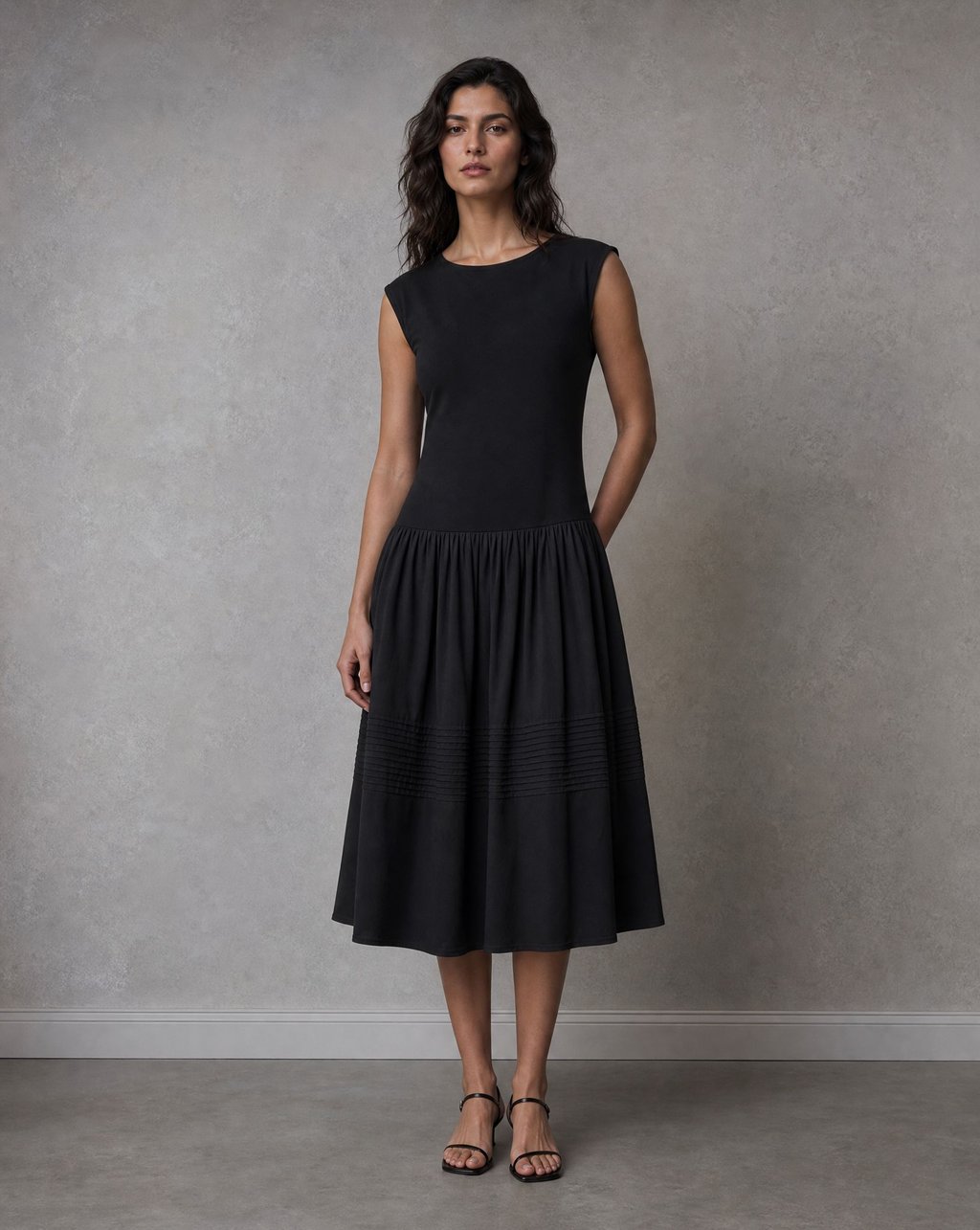 A model wearing a black pleated midi dress, photographed full length against a grey wall in soft even light with sandals visible