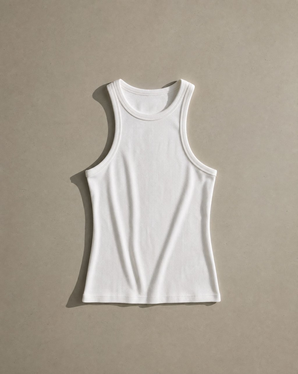 A white ribbed tank top laid flat on a warm grey surface with a strong directional shadow falling to the left