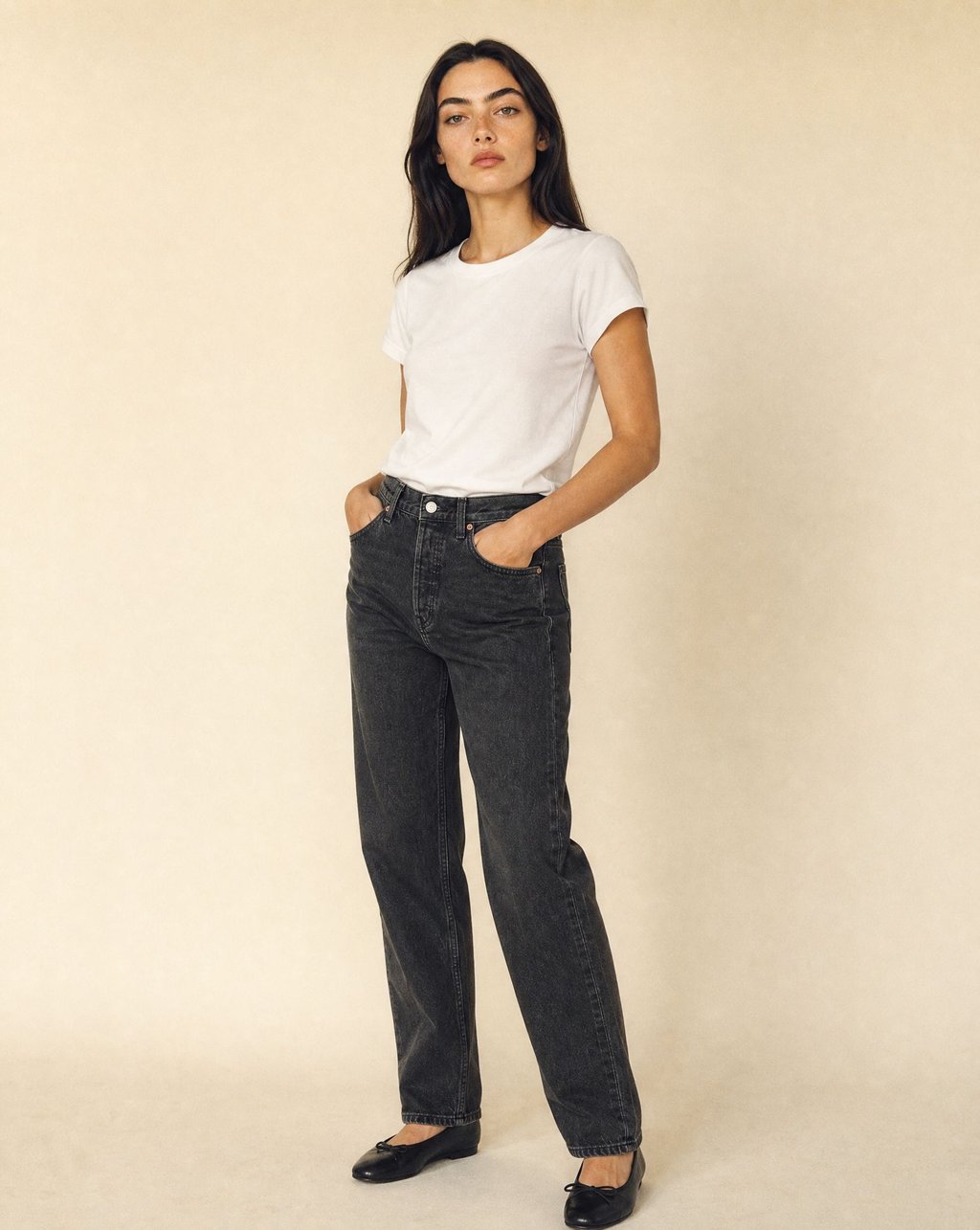 A woman in a white tee wearing washed black denim jeans against a warm cream backdrop