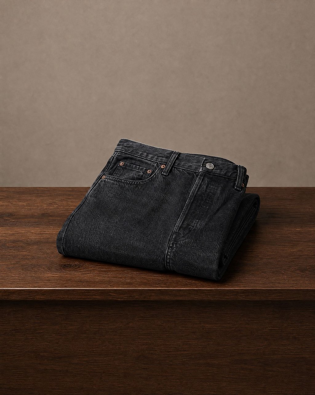 Black denim jeans folded on a dark wood ledge under soft light, with the denim weave, copper rivets and stitching legible and empty space around them