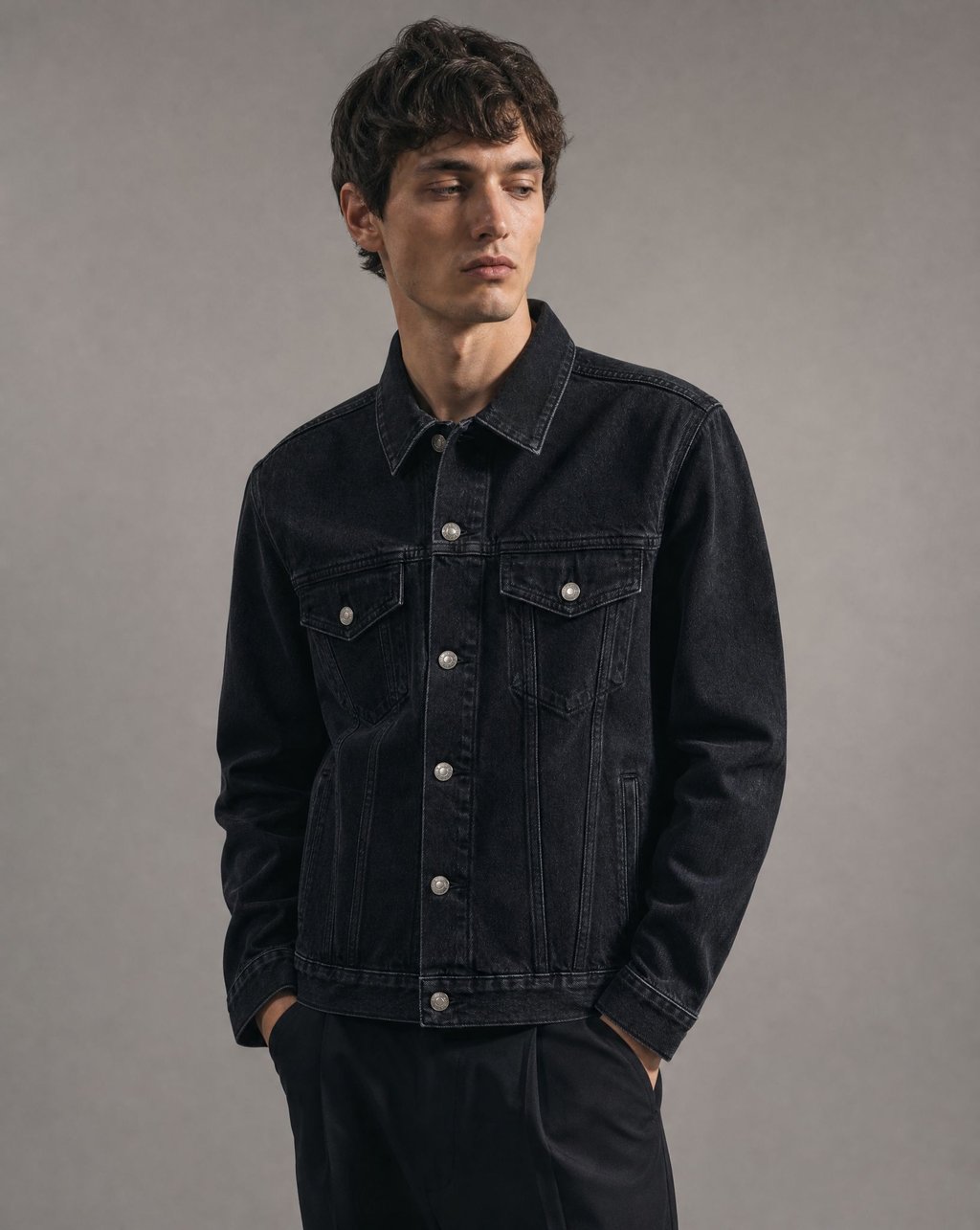 Editorial studio portrait of a model in a black denim jacket against a grey backdrop