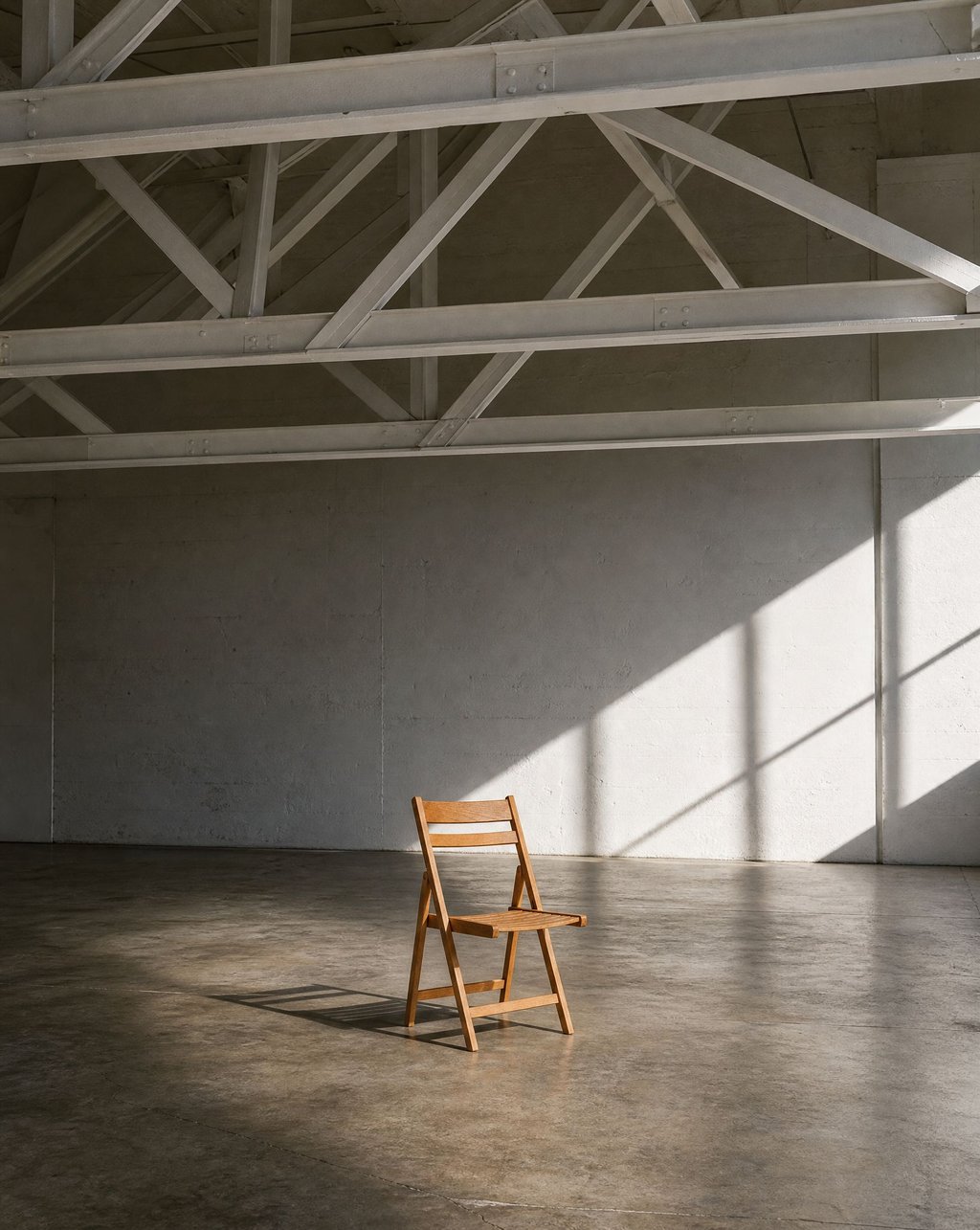 A light wooden folding chair in a bare concrete room cut by hard angular beams of light