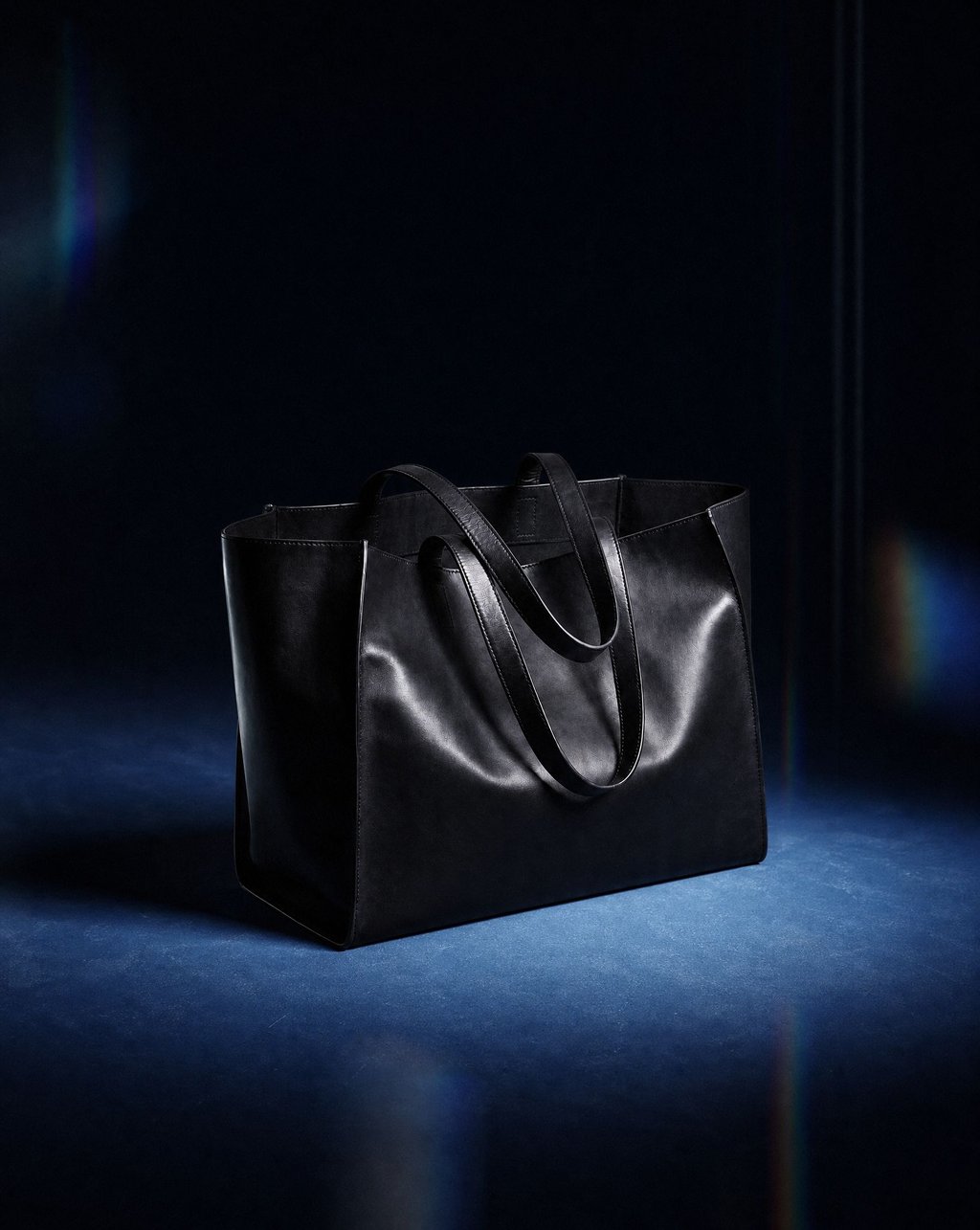 A black leather tote bag on a blue floor against a dark background, lit by a narrow spotlight with faint lens flares