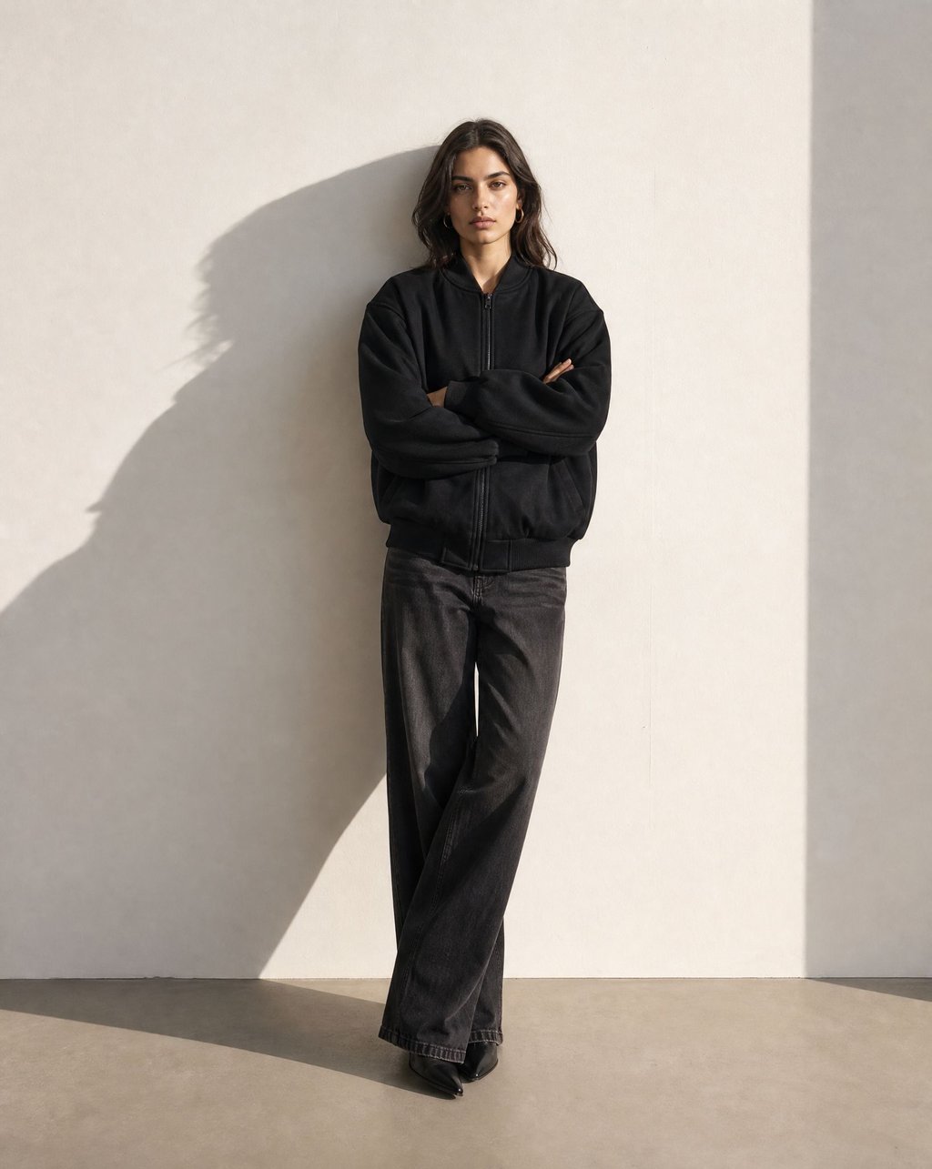 A model wearing a black bomber jacket against a pale wall, with a strong directional shadow cast across the wall