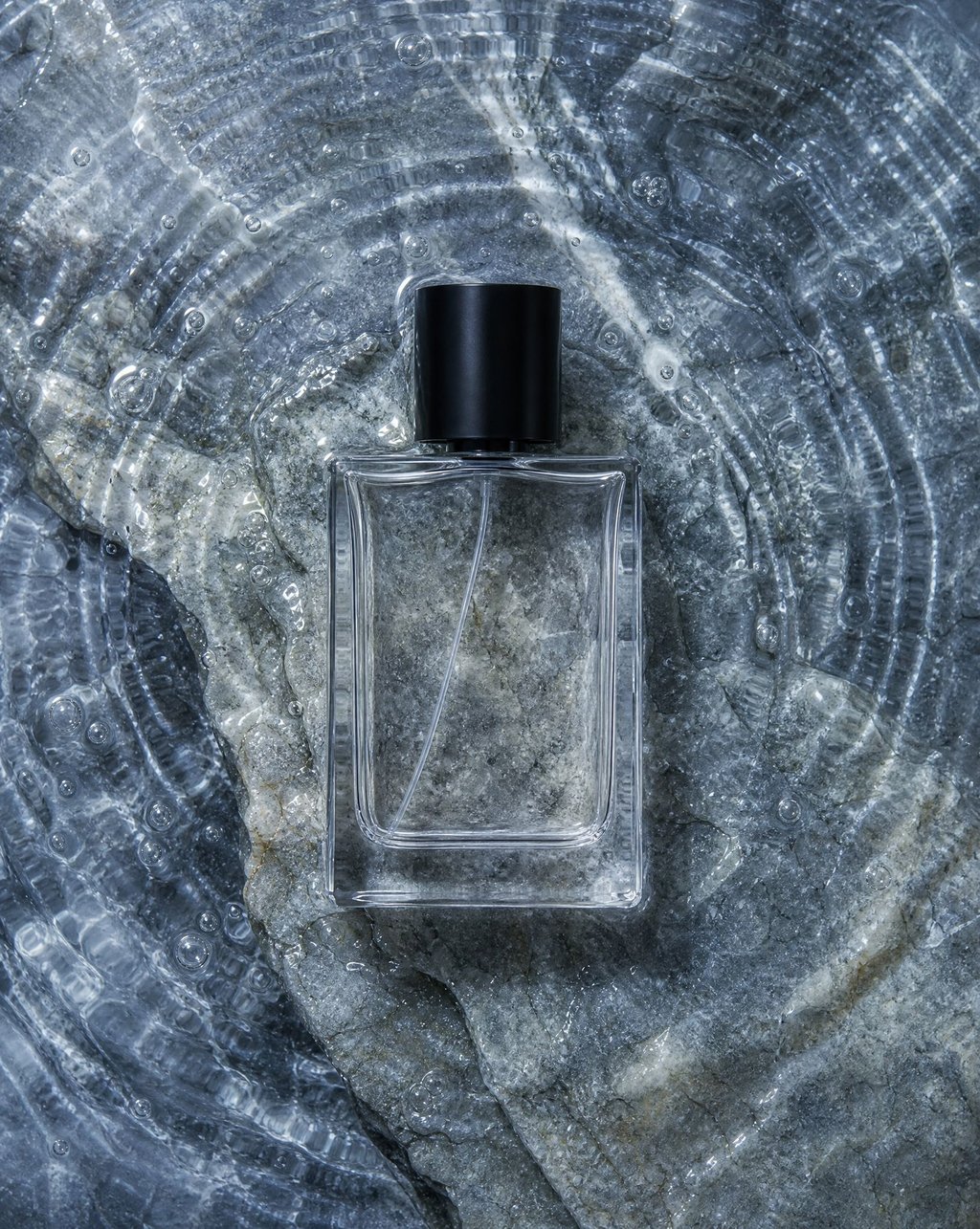 Rectangular glass perfume bottle lying on wet glacial stone with water ripples catching overhead light