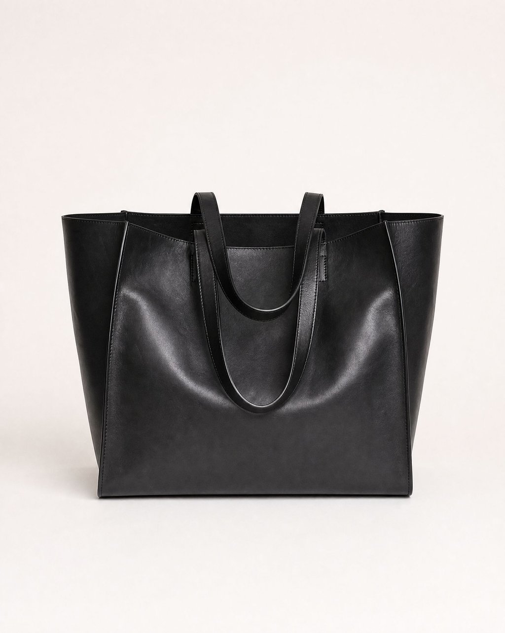 A black leather tote bag on a plain warm-white background under flat even light