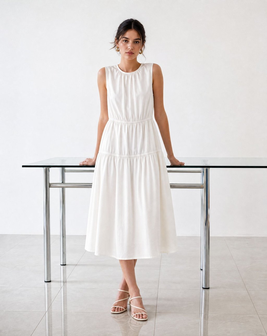 A model in a cream tiered midi dress leaning against a glass table in even neutral light, the fabric drape clearly visible