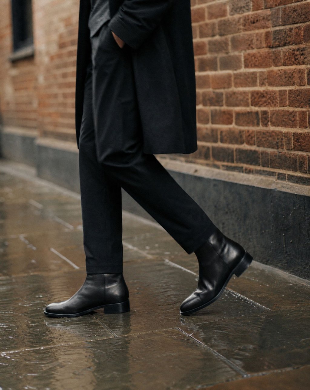 Black leather ankle boots worn on a wet sidewalk beside a plain brick wall