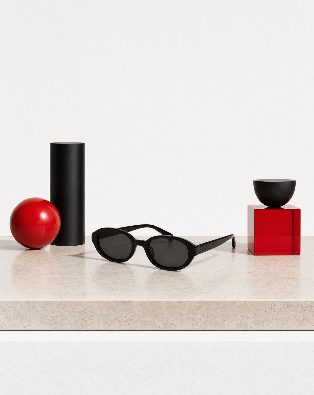 Black oval sunglasses on a pale stone shelf with a red sphere, a black cylinder and a red glass cube behind them