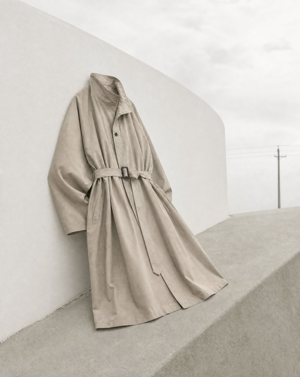 A greige belted trench coat leaning against a curved white wall under diffused overcast light