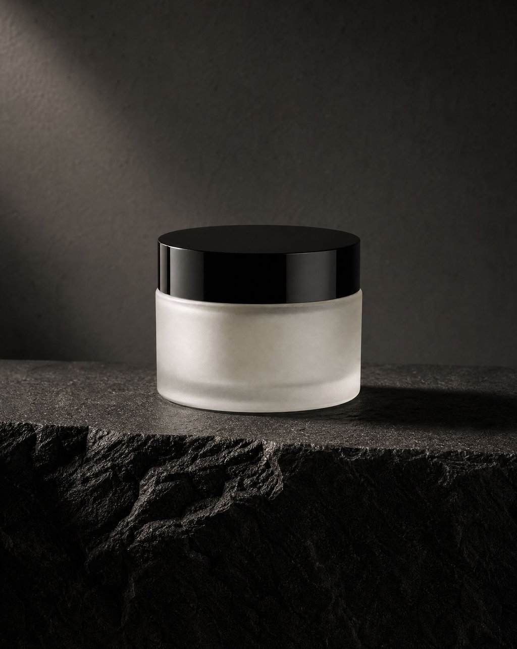 A frosted glass skincare jar with a black lid standing on a dark basalt stone ledge under dramatic directional light