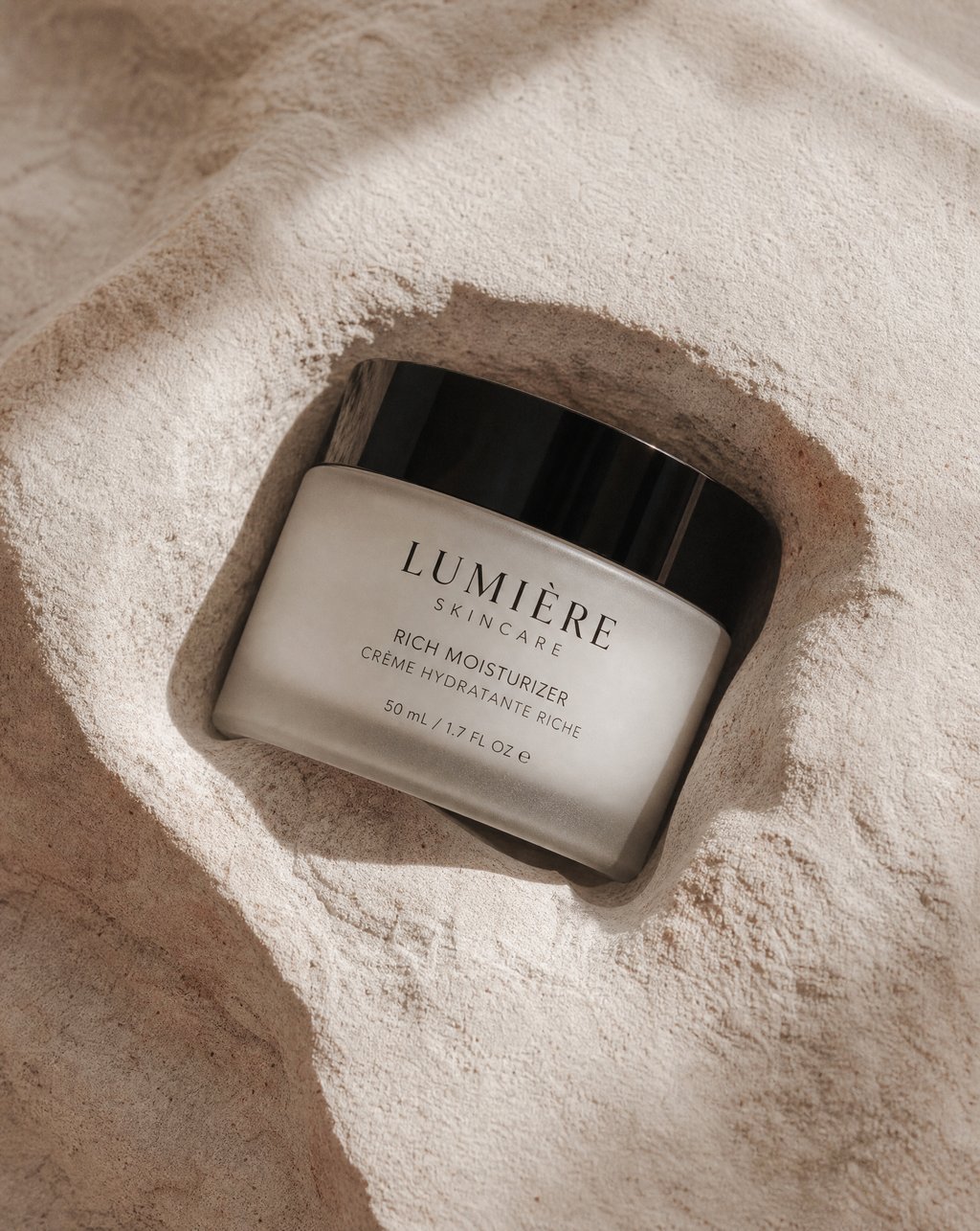 A frosted glass moisturizer jar with a black lid resting in a hollow of pale textured stone, lit by soft window light with the label fully legible