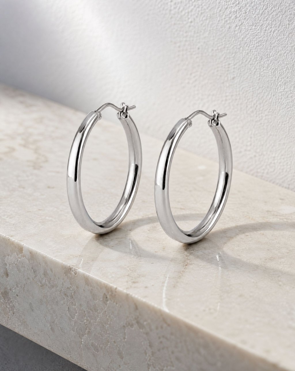 Pair of polished silver hoop earrings standing on a pale marble ledge against a textured white wall in cool diffused light