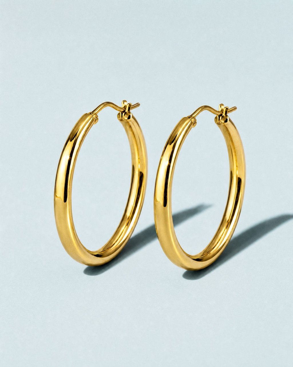 A pair of polished gold hoop earrings standing upright on a pale blue surface under hard direct flash