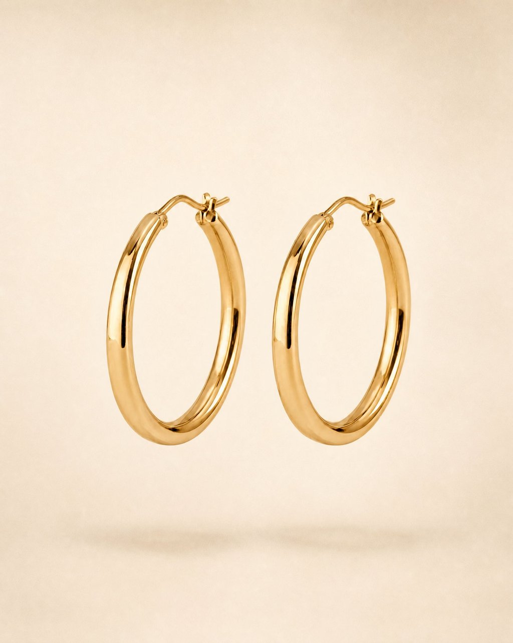 Pair of polished hoop earrings floating over a cream studio backdrop with a soft shadow