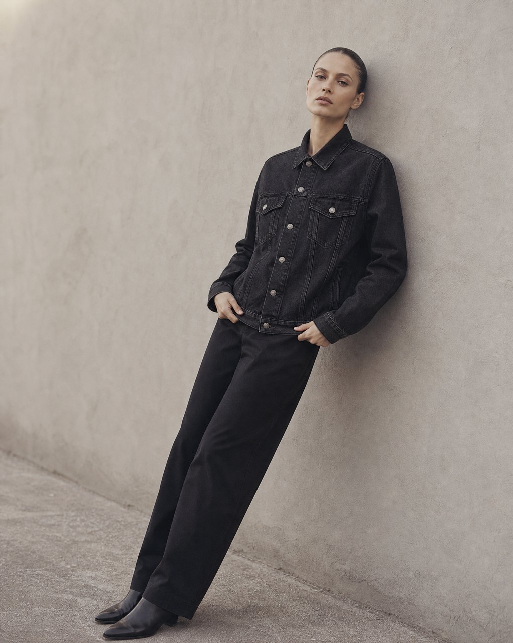 A model in a black denim jacket leaning against a plain brushed concrete wall in soft overcast light, a clean lookbook-style frame