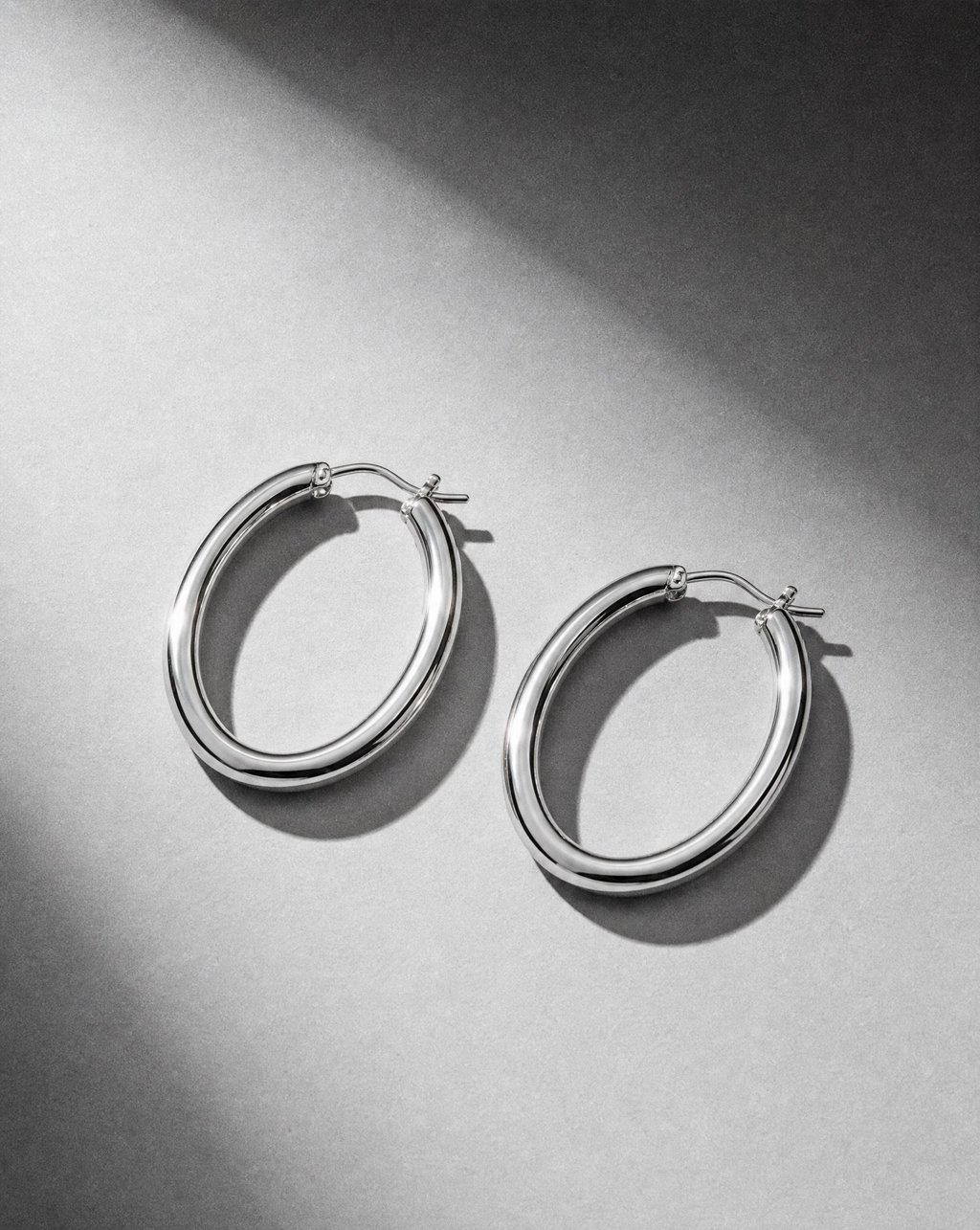 A pair of polished silver hoop earrings on a grey surface under hard directional light, with crisp highlights along each hoop and defined shadows beneath