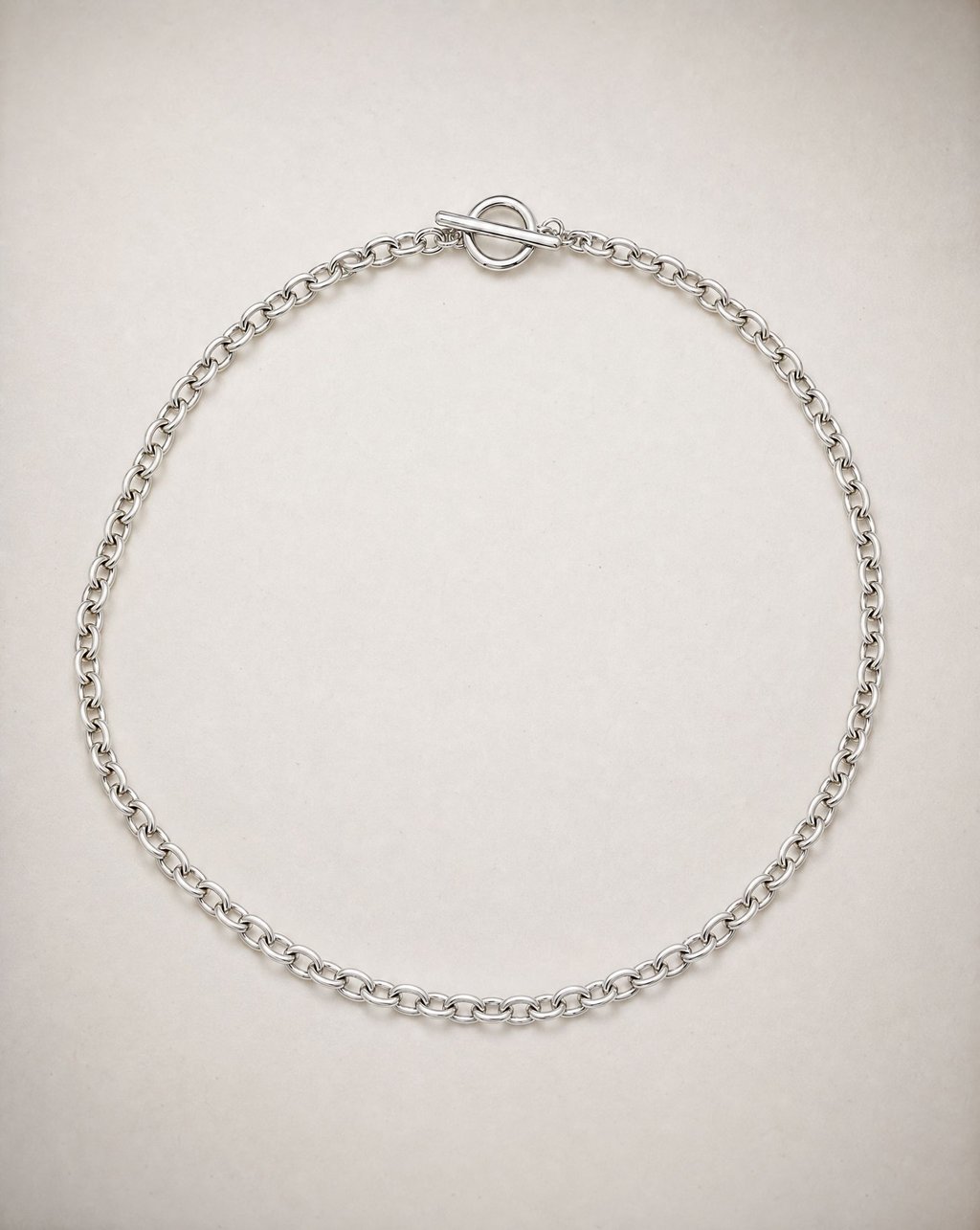 A silver link chain necklace laid flat in a circle on a pale surface under soft overhead light, with the toggle clasp visible at the top