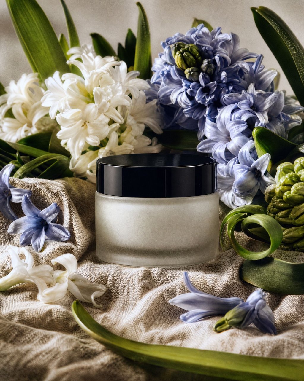 Frosted glass skincare jar with a black lid standing on crumpled linen among white and blue hyacinth flowers
