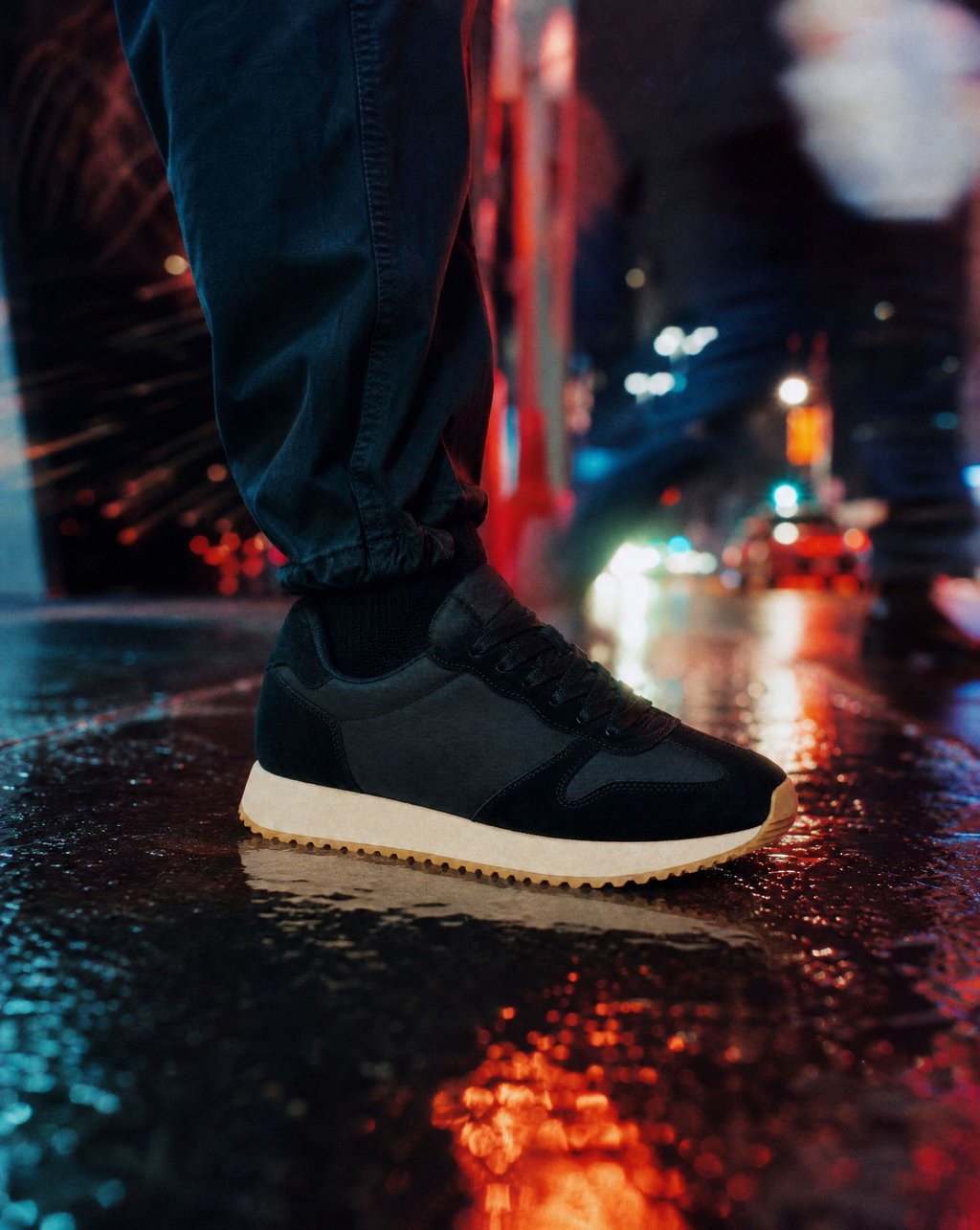 A black trainer with a cream sole on wet city pavement reflecting red and blue neon light at night