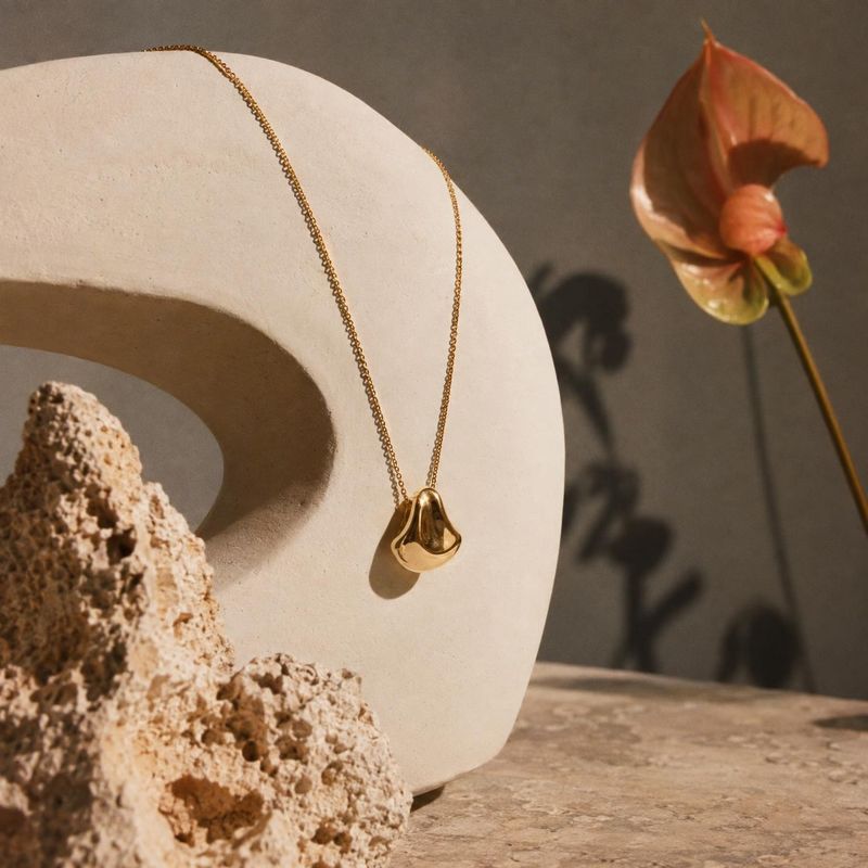 gold pendant necklace close-up detail product photo: A sculptural gold pendant necklace drapes over a pale stone form beside a textured rock and an orange flow…
