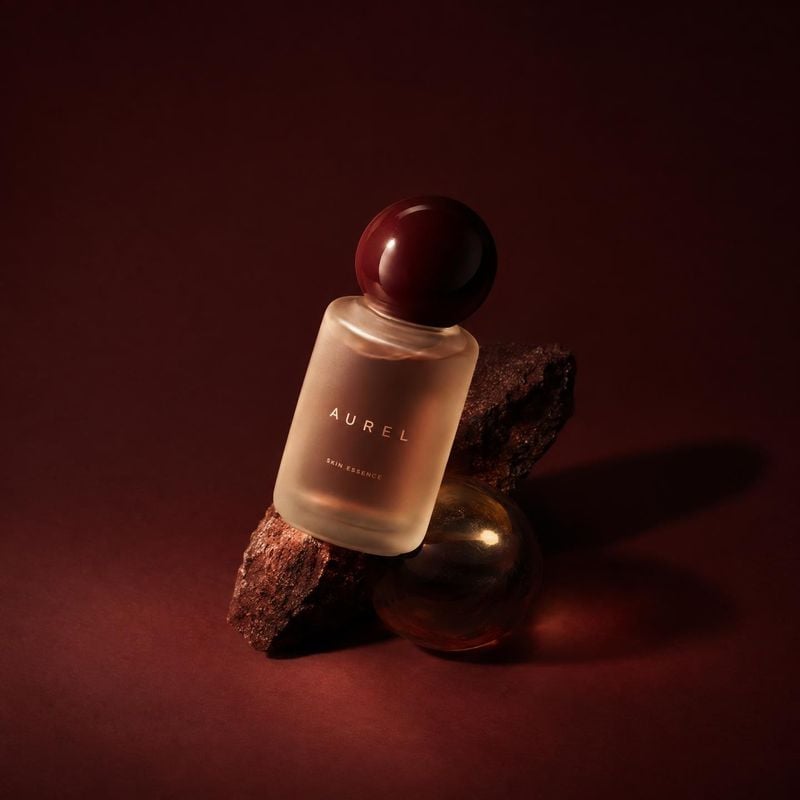 skin essence still life product photo: A frosted bottle of skin essence rests among dark stone and glass props against a deep burgundy studio backdrop, lit wit…