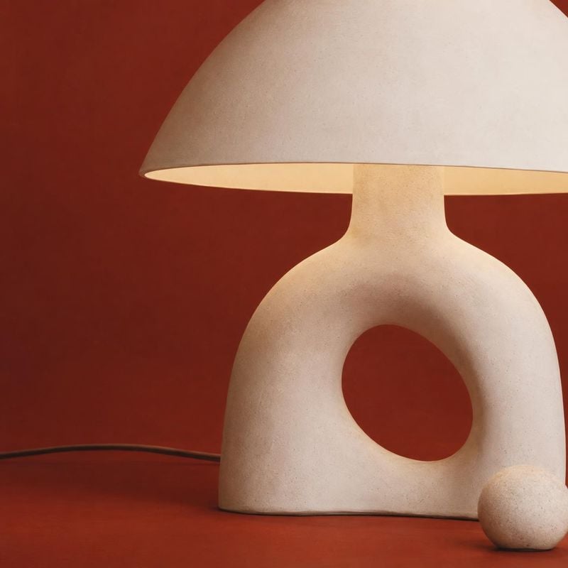 ceramic table lamp still life product photo: A sculptural cream ceramic table lamp with a rounded open-form base sits against a terracotta-red studio backdrop,…