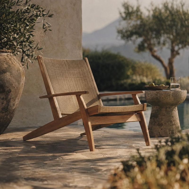 wooden lounge chair lifestyle scene product photo: A woven wooden lounge chair sits on a stone patio beside an olive tree and a small stone table, photographed…