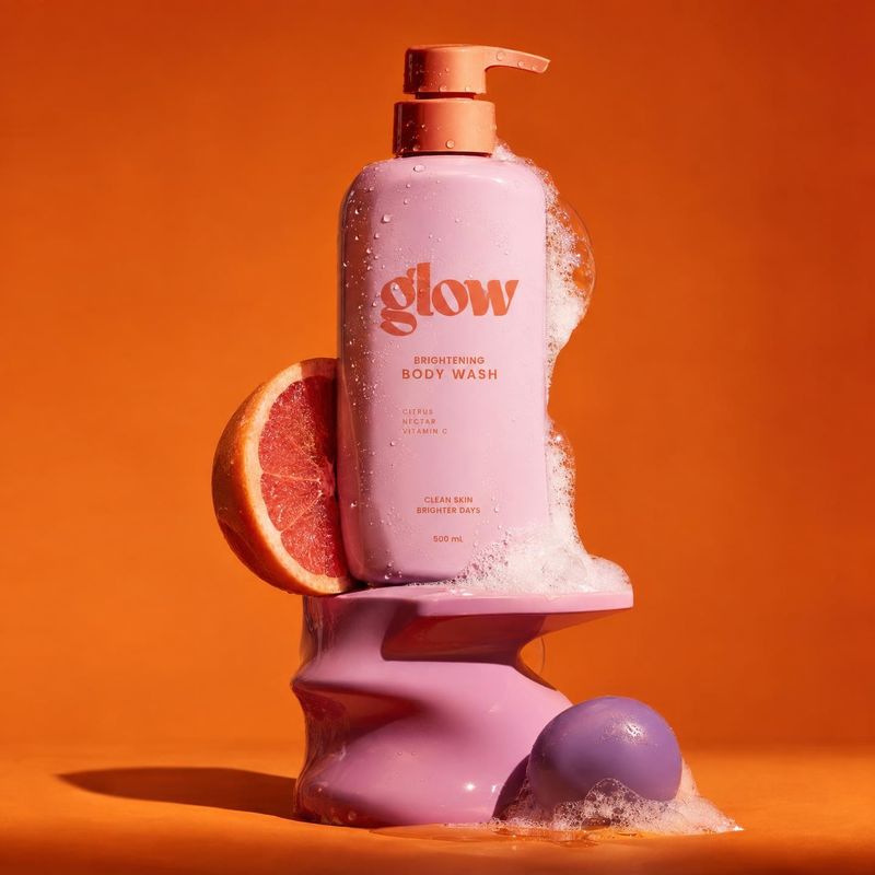 body wash ingredients product photo: A pink body-wash pump bottle beaded with water and foam sits on a sculptural pink pedestal beside a cut grapefruit, agains…