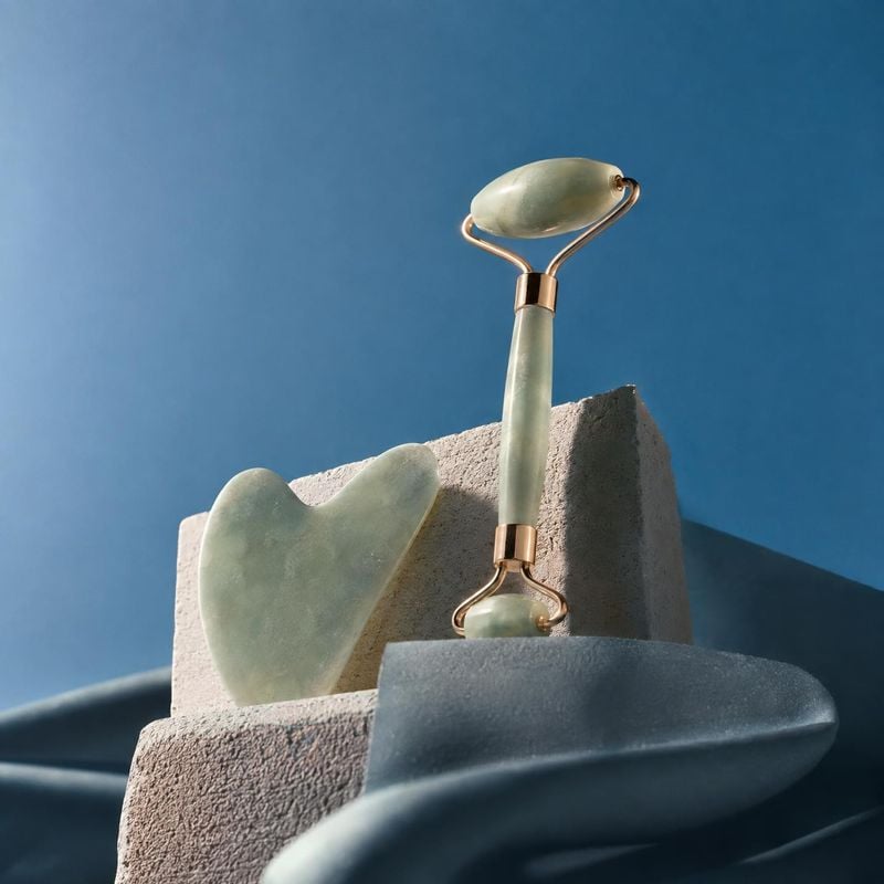 jade facial roller and gua sha tool still life product photo: Jade facial massage tools are arranged on sculptural stone blocks against a clear blue backdrop,…