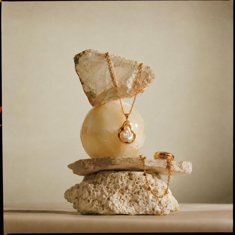gold jewelry set close-up detail product photo: A gold pendant necklace and ring are artfully arranged on pale stone and a polished sphere against a warm neutr…