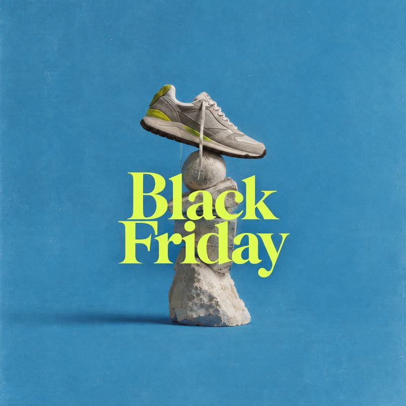 sneaker fashion product photo: A gray sneaker balances atop a sculptural stack of stones against a textured blue studio backdrop, with oversized neon-yellow Bl…