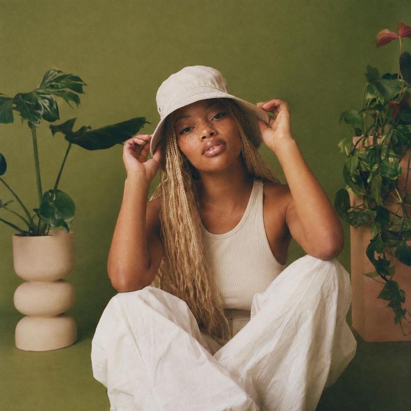 women's outfit and bucket hat look book product photo: A seated model in a cream bucket hat, ribbed tank and flowing white trousers poses among potted plants a…