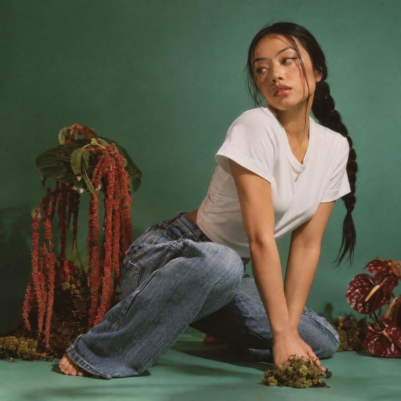 casual outfit with wide-leg jeans look book product photo: A woman in a white T-shirt and loose blue jeans poses barefoot among leafy plants against a deep gre…