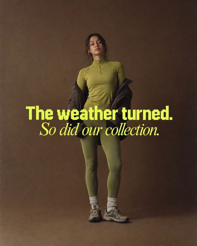 women's athletic outfit fashion product photo: A woman poses in an olive-green athletic outfit with a dark jacket draped around her arms against a warm brown s…