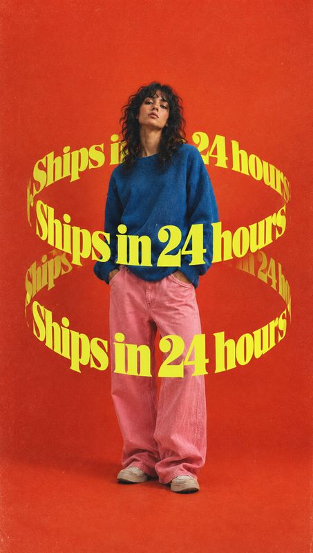 casual outfit model try-on product photo: A model in a blue sweater and pink trousers stands against a vivid red studio backdrop, encircled by bold yellow “Shi…