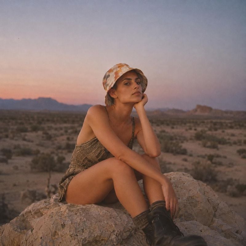 fashion outfit look book product photo: A woman in a patterned bucket hat, textured outfit, and boots sits on a rock in a desert landscape at dusk, with soft p…