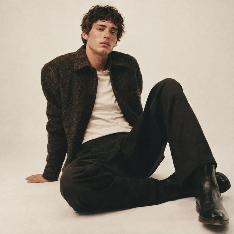 men's outfit look book product photo: A casually seated male model wears a dark textured jacket, light shirt, black trousers, and boots against a warm off-whit…