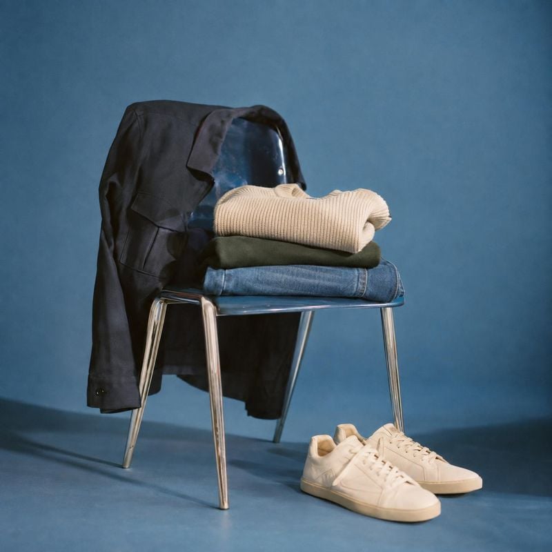 casual clothing and sneakers still life product photo: A neatly styled outfit of folded knitwear, denim, a draped jacket and pale sneakers rests on a metal cha…