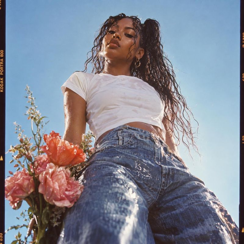 denim jeans look book product photo: A model in a white crop top and wide-leg denim jeans poses from a dramatic low angle among pink flowers beneath a bright b…