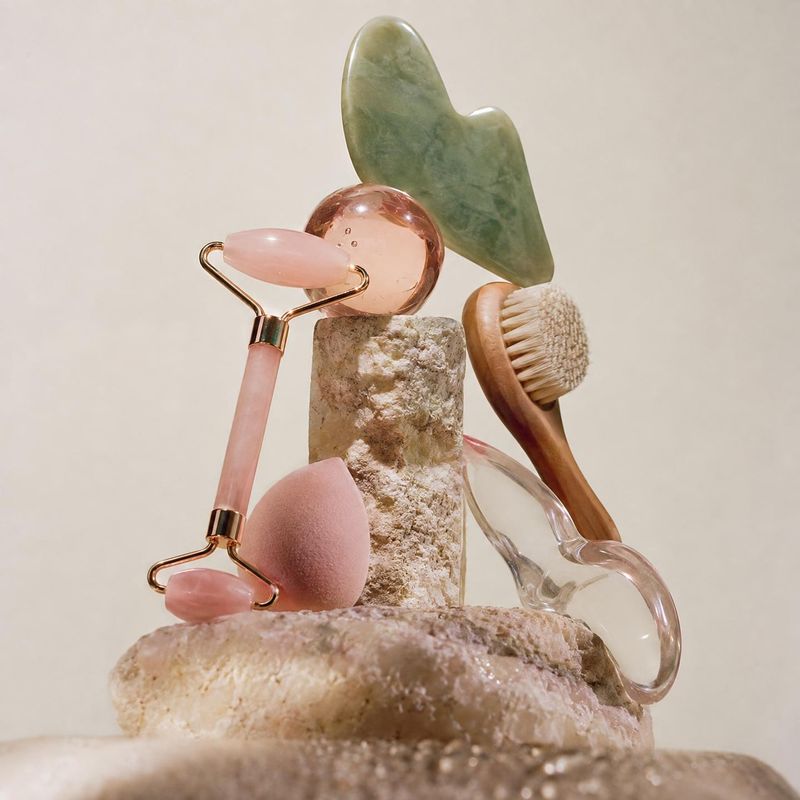 facial massage tools close-up detail product photo: A pink facial roller, stone gua sha tools, a brush, and a sponge are artfully arranged on textured stone ag…