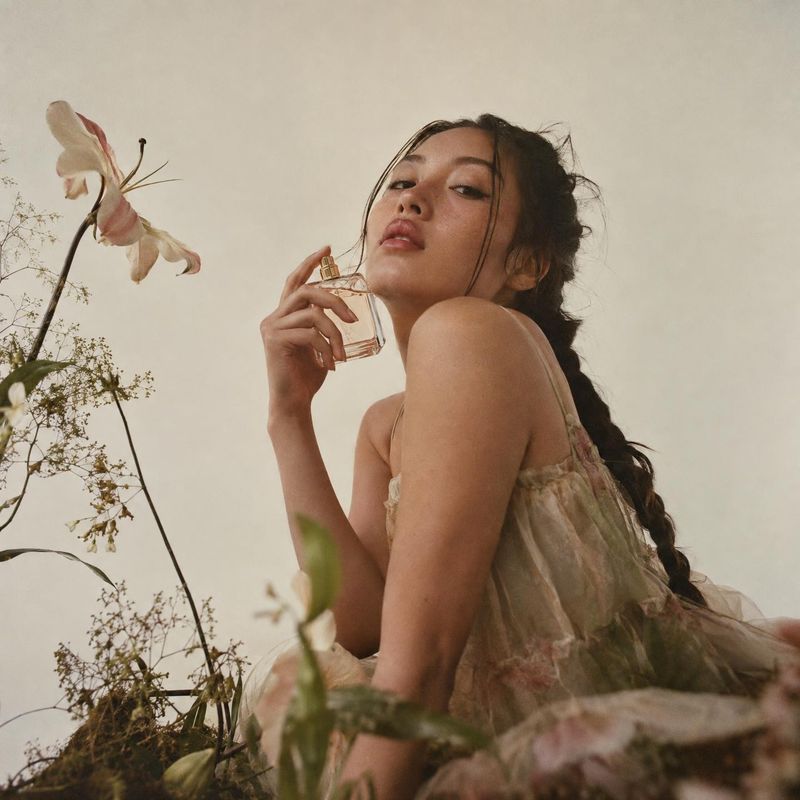 perfume look book product photo: A woman in a romantic floral dress holds a clear perfume bottle near her face amid delicate flowers, in soft warm light with a…